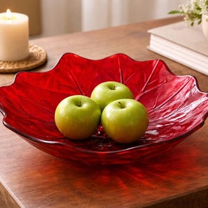 May include: A red, leaf-shaped glass bowl holds three green apples. A lit candle sits on a woven coaster to the left. A small vase with white flowers and a book are in the background, all on a wooden table.