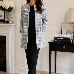 May include: A black and white tweed coat with a round collar, worn over a navy top and black pants. The coat has long sleeves and pockets. The outfit is completed with black heels. The setting is a well-lit room with a fireplace and a side table.