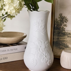 May include: A white ceramic vase with a scalloped rim and a raised floral relief pattern featuring leaves and blossoms on the exterior surface. The vase has a tapered neck and a rounded base, holding a bouquet of white hydrangea flowers.