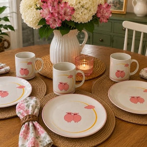 May include: A dinnerware set featuring three white ceramic mugs and three white ceramic plates, each decorated with a graphic of two pink sheep standing together under a yellow crescent moon and a small pink cloud, arranged on woven placemats.