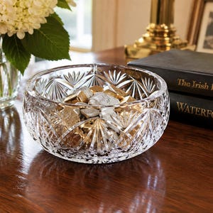 May include: A clear, round crystal bowl featuring intricate fan-shaped and diamond-cut patterns across its exterior surface. The bowl is filled with small, individually wrapped gold and silver candies, showcasing a reflective and faceted glass design.