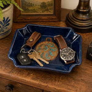 May include: A deep blue ceramic catchall tray containing a brown leather keychain with metal keys, a round decorative medallion with a teal and brown floral pattern, and a wristwatch with a brown leather strap and a black analog face.