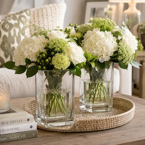 May include: Two clear square glass vases filled with white hydrangeas, white roses, green carnations, and small green berries. The floral arrangements are displayed on a round woven tray next to a stack of three books with visible text.