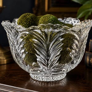 Peut inclure: Un bol en verre transparent avec un motif de feuilles d&eacute;taill&eacute;, rempli de boules de mousse vertes. Le bol a un bord festonn&eacute; et repose sur une surface en bois sombre. Un bougeoir dor&eacute; et des livres sont en arri&egrave;re-plan.