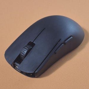 Könnte beinhalten: Eine mattschwarze kabellose Computermaus mit einer glatten, ergonomischen Schale. Sie verfügt über ein zentrales Scrollrad mit texturiertem Griff, zwei Hauptklicktasten und zwei zusätzliche Seitentasten an der linken Kante des Geräts.