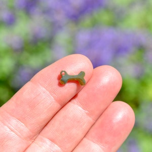 May include: A small, gold-colored dog bone charm with a loop for attaching to a necklace or bracelet. The charm is held between two fingers against a blurred background of green and purple foliage.