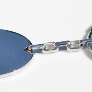 Puede incluir: Un llavero plateado con una etiqueta ovalada en blanco. La etiqueta est&aacute; conectada a una cadena de peque&ntilde;os eslabones rectangulares, que conduce a un gran llavero circular. El llavero est&aacute; hecho de un metal brillante y reflectante.