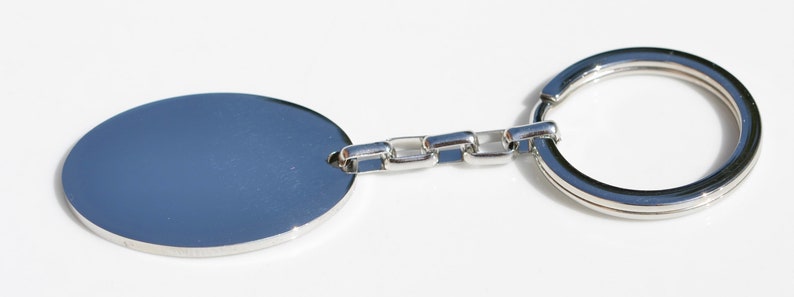 Puede incluir: Un llavero plateado con una etiqueta ovalada en blanco. La etiqueta est&aacute; conectada a una cadena de peque&ntilde;os eslabones rectangulares, que conduce a un gran llavero circular. El llavero est&aacute; hecho de un metal brillante y reflectante.