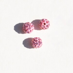 May include: Three pink round beads with a sparkly finish. Each bead is covered in small pink rhinestones.