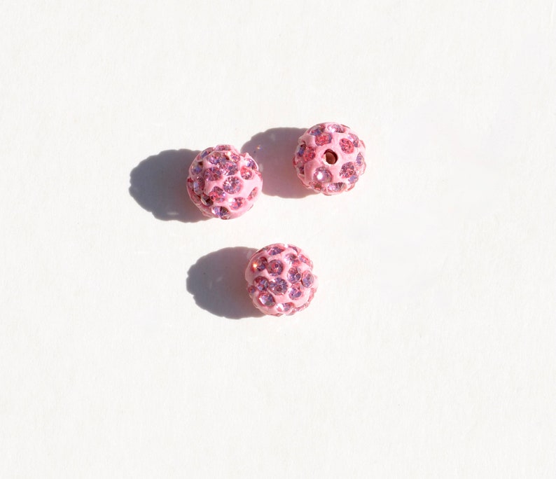May include: Three pink round beads with a sparkly finish. Each bead is covered in small pink rhinestones.