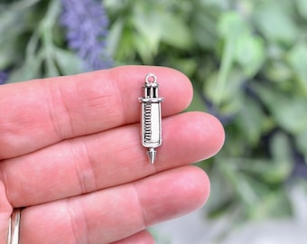 May include: Silver-toned metal syringe charm with a detailed design. The charm is about 1 inch long and has a loop at the top for attaching to a necklace or bracelet.