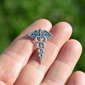 May include: Silver-toned medical caduceus charm with a snake wrapped around a staff and wings. The charm is on a person's hand.