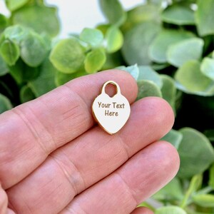 May include: A small, gold-colored heart-shaped charm featuring a smooth, polished surface with the engraved text "Your Text Here" centered on the front. The charm includes a small circular loop at the top for attachment to jewelry or accessories.