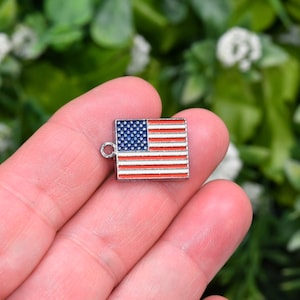 May include: A small silver charm in the shape of the American flag. The flag is red, white, and blue with a silver loop for attaching to a necklace or bracelet.