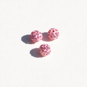 May include: Three pink round beads with a sparkly finish. Each bead is covered in small pink rhinestones.