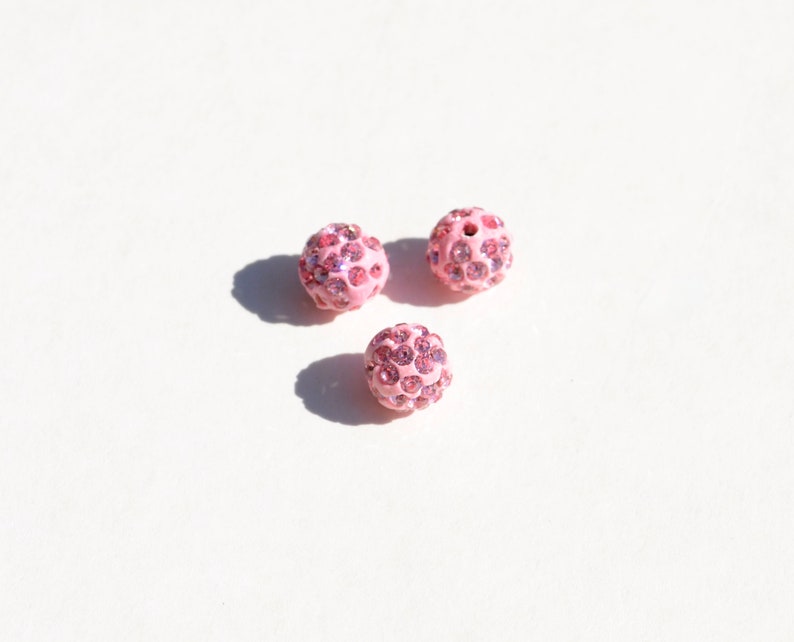 May include: Three pink round beads with a sparkly finish. Each bead is covered in small pink rhinestones.