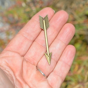 May include: A gold-toned arrow charm with a detailed arrowhead and fletching. The charm is held in a hand, showcasing its size and detail.