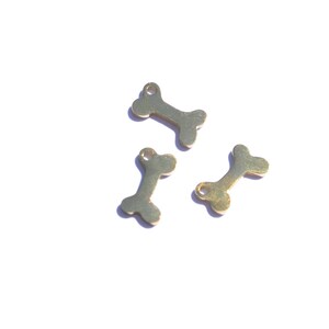 May include: Three small, gold-colored bone charms are scattered on a white surface. Each charm has a small hole at one end, suggesting they are designed for jewelry making or crafting. The bone shape is clearly defined.
