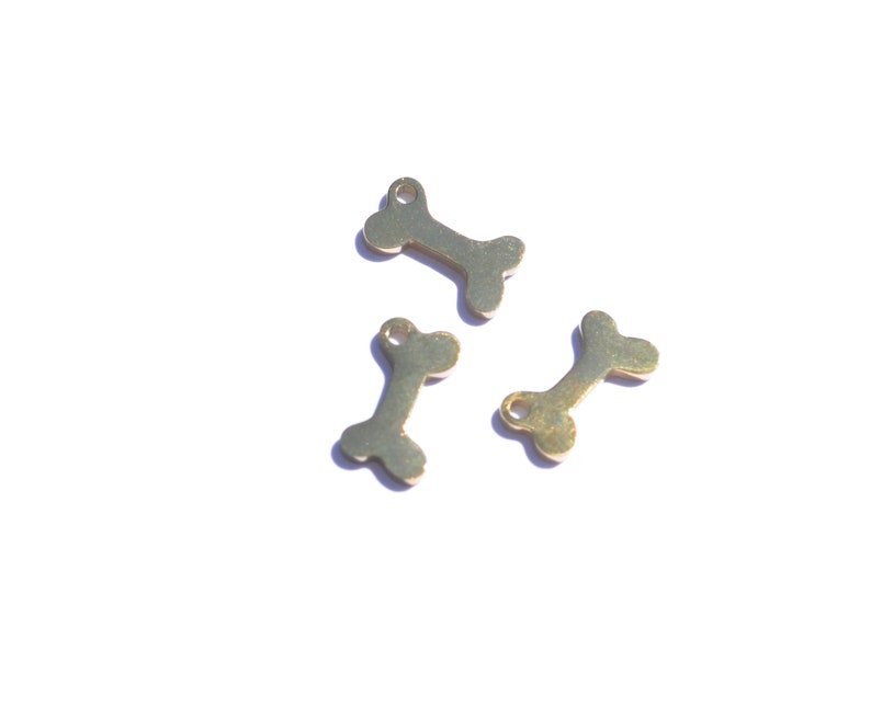 May include: Three small, gold-colored bone charms are scattered on a white surface. Each charm has a small hole at one end, suggesting they are designed for jewelry making or crafting. The bone shape is clearly defined.