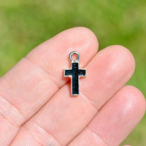May include: A small, silver-toned cross pendant with a black enamel center. The cross has a loop at the top for attaching to a chain or cord. The background is a soft, blurred green.
