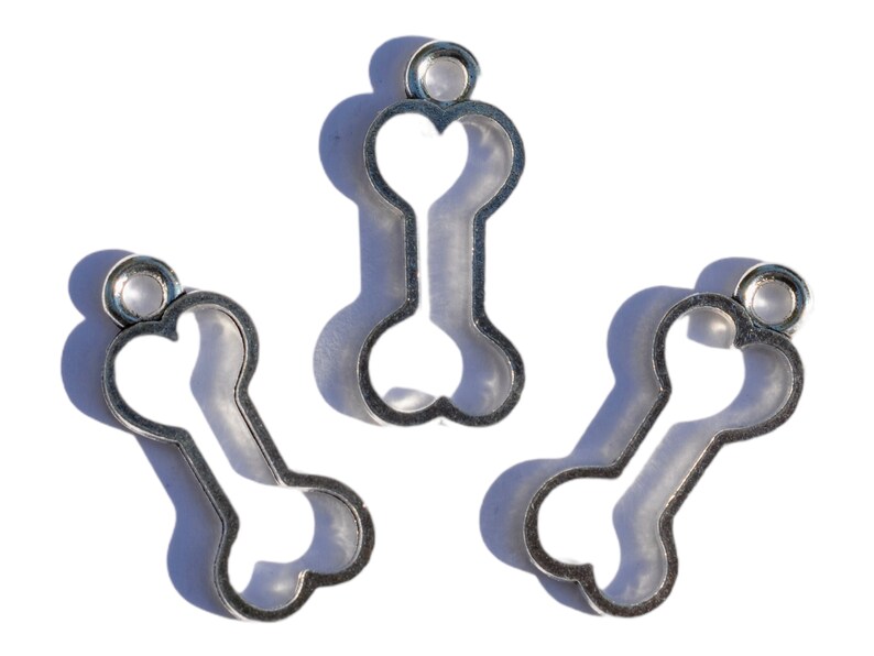 K&ouml;nnte beinhalten: Drei silberne Metall-Anh&auml;nger in Form von Hunde-Knochen. Jeder Anh&auml;nger hat oben eine Schlaufe zum Anbringen an eine Halskette oder ein Armband.