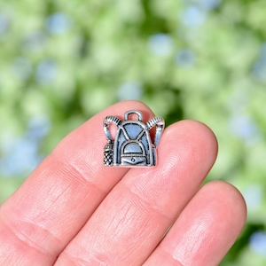 May include: Silver charm in the shape of a backpack with a water bottle attached. The charm is small and detailed.