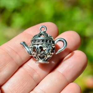 May include: Silver charm in the shape of a teapot with a heart design. The teapot has a detailed, intricate design with a heart pattern.