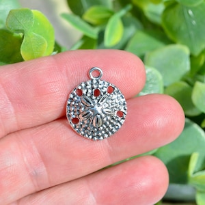 May include: Silver charm in the shape of a sand dollar with a detailed design. The charm has a loop at the top for attaching to a necklace or bracelet.