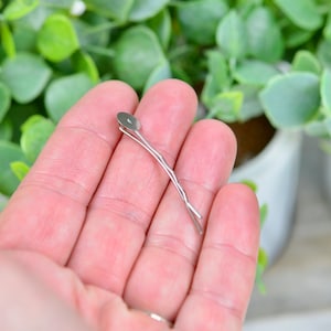 May include: A silver hair clip with a round, flat top. The clip is held in a hand.