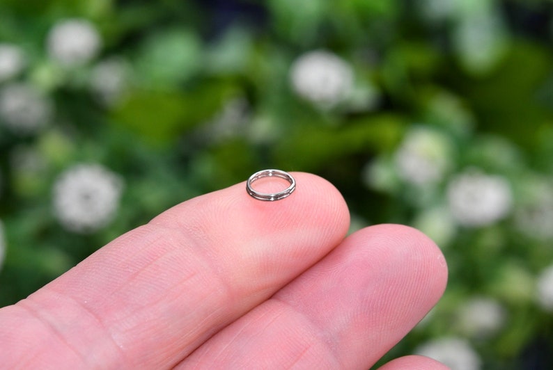May include: A small, silver ring, likely a nose ring, is held between two fingers.