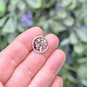 May include: Silver charm in the shape of a tree of life. The charm is round and has a detailed design of a tree with branches and leaves.