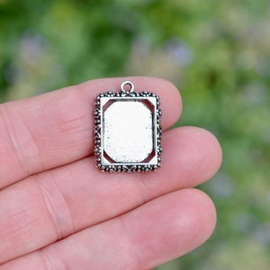 May include: A rectangular silver-toned pendant with a decorative beaded border and a small loop at the top. The center of the pendant is a recessed area, likely for adding a design or image. The pendant is held in a hand against a blurred green background.