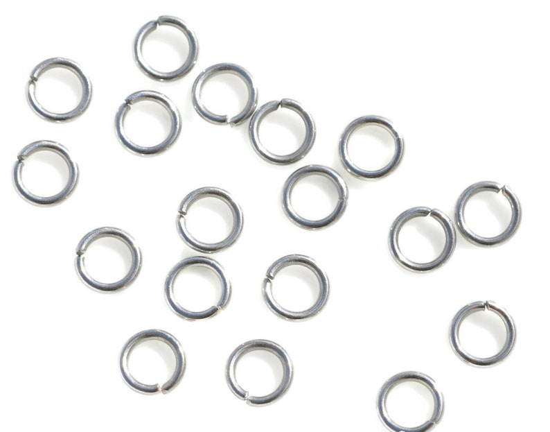 May include: A collection of silver jump rings, scattered on a white background. The jump rings are small and circular, with a gap in the ring.