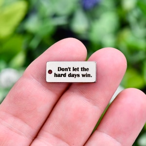 May include: Silver charm with a rectangular shape and a hole for attaching to a necklace or bracelet. The charm has the text "Don't let the hard days win." engraved on it.