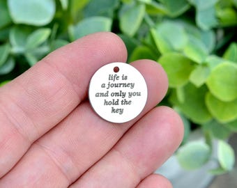 May include: A small, circular silver-toned metal charm featuring a hole at the top for attachment. The surface is engraved with the black text: life is a journey and only you hold the key. The charm has a smooth, polished finish.