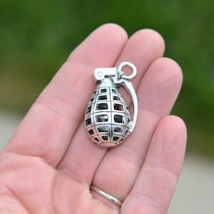 May include: Silver charm in the shape of a grenade. The grenade is detailed with a grid pattern and has a loop at the top for attaching to a chain or bracelet.