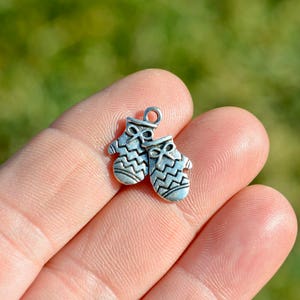 May include: Two small, silver-colored mitten charms. Each charm has a loop at the top for attaching to a chain or bracelet. The mittens have a zig-zag pattern and a bow detail.