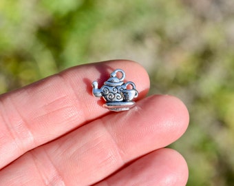 May include: Silver charm in the shape of a teapot and cup. The teapot has a decorative design and the cup has a handle.