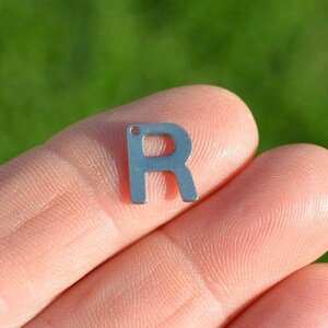 May include: A silver metal letter R charm. The charm is held between two fingers.