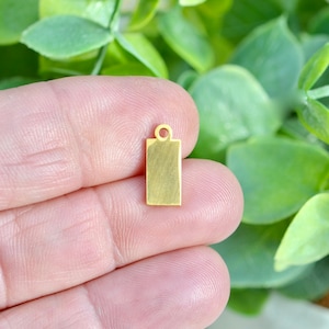 May include: A gold-colored metal tag with a loop at the top. The tag is rectangular and has a brushed finish.