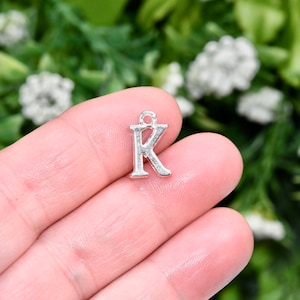 May include: Silver charm in the shape of the letter K. The charm is shown in a hand against a green background.