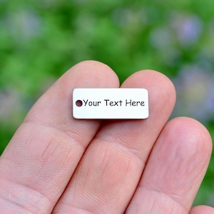 May include: A small, rectangular, silver-colored metal charm with rounded corners and a single circular hole on the left side. The surface of the charm features the black engraved text "Your Text Here" centered on the front face of the piece.