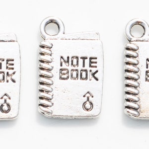 K&ouml;nnte beinhalten: Drei silberne Anh&auml;nger in Form von Notizb&uuml;chern mit der Gravur "NOTE BOOK". Jeder Anh&auml;nger hat oben eine &Ouml;se zum Anbringen an einer Halskette oder einem Armband.