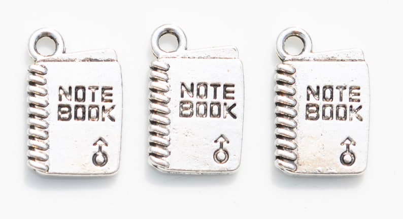 K&ouml;nnte beinhalten: Drei silberne Anh&auml;nger in Form von Notizb&uuml;chern mit der Gravur "NOTE BOOK". Jeder Anh&auml;nger hat oben eine &Ouml;se zum Anbringen an einer Halskette oder einem Armband.