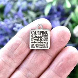 May include: A square silver-toned metal charm featuring a small loop at the top. The surface is engraved with a camper graphic and the words CAMPING, FRIENDS, S'MORES, STARS, FAMILY, ADVENTURE, CAMP COFFEE, FISHING, RELAX, UNWIND, and NATURE.