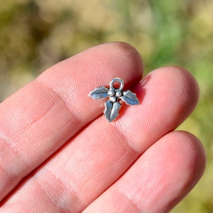 May include: Silver charm in the shape of a holly leaf with three red berries. The charm has a loop at the top for attaching to a necklace or bracelet.