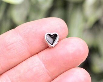 May include: A small heart-shaped charm featuring a polished silver-toned metal border surrounding a smooth, glossy black center inlay. The piece is compact and symmetrical, showcasing a clean, defined outline that highlights the contrast between the dark interior and the metallic frame.
