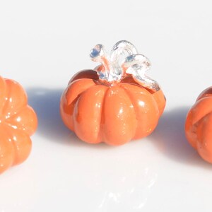 May include: Three small orange pumpkin charms with silver stems. The pumpkins are smooth and have a slightly rounded shape.
