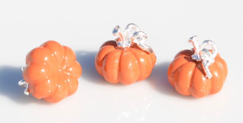 May include: Three small orange pumpkin charms with silver stems. The pumpkins are smooth and have a slightly rounded shape.