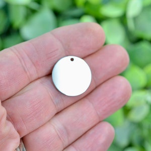 May include: A small, circular, silver-colored metal disc charm with a smooth, reflective surface. The disc features a single, centered circular hole near the top edge, designed for attachment to jewelry or other decorative items.